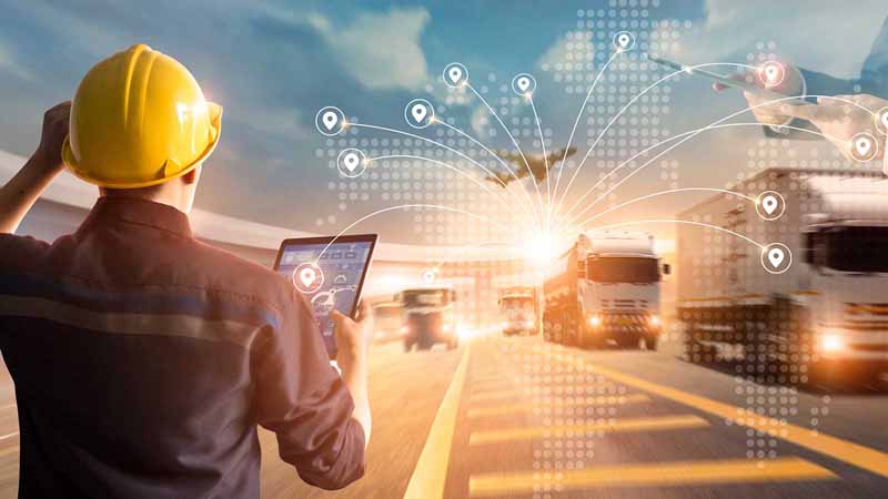 A worker in a hard hat holding a tablet and watching supply trucks.